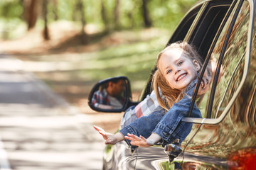 Obraz premium Cute cheerful little caucasian girl sitting inside the car, looking out the window and smiling. Family road trip