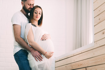Young pregnant couple near the window of the house, on the background of a brick white wall and a wooden window sill