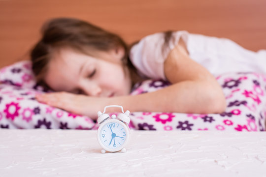 White alarm clock stands on the table on the background of a sleeping girl