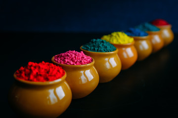 Line of colorful Holi powder in cups closeup. Bright colours for Indian holi festival in clay pots. Selective focus. Black background. Happy Holi greeting card