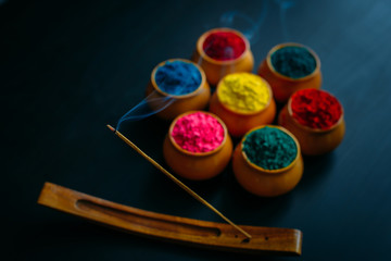 Burning and fuming aroma stick against circle of Holi powder in clay cups. Black background.  Happy...