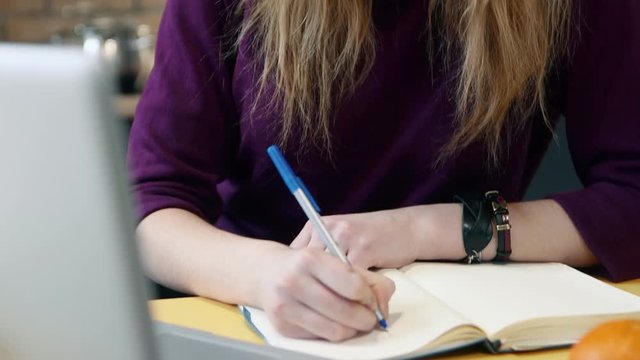 Woman learns on a computer and and writes notes in notepad in the kitchen at home.