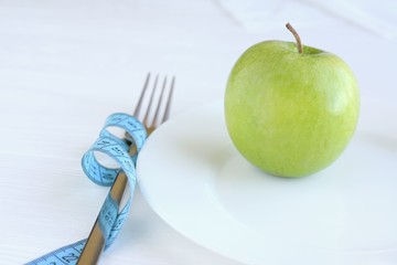 Juicy organic green apple on white plate with selective focus and blurred fork with blue measure tape on neutral background. Diet concept with healthy vegetarian food for weight loss