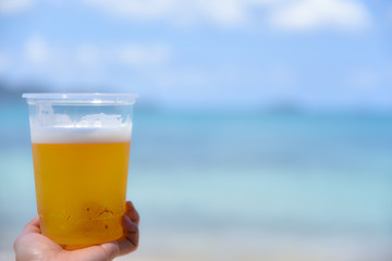 Woman hand holding a glass of beer on the beach with blurred sea and blue sky background. Summer day concept. Vacation holidays background wallpaper.
