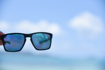 Man hand holding sunglasses on the beach with blurred sea and blue sky background. Summer day concept. Vacation holidays background wallpaper.