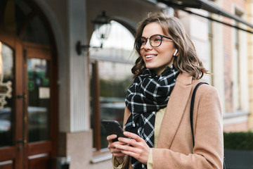 Young woman outdoors walking by street using mobile phone listening music with earphones.