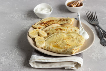 Thin pancakes or crepes with natural yogurt, honey and banana on a plate. Light concrete background. Copy space.