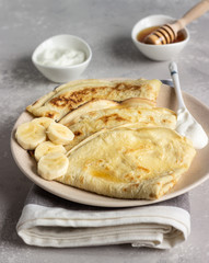 Thin pancakes or crepes with natural yogurt, honey and banana on a plate. Light concrete background. Copy space.