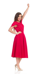 Woman In Elegant Red Dress And Gold High Heels Is Standing With Arm Raised, Looking At Camera And Smiling