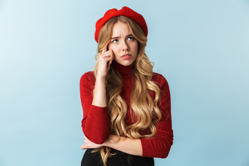 Portrait of discontent blond woman 20s wearing red beret looking aside while standing, isolated over blue background in studio