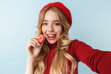 Image of european blond woman 20s wearing red beret eating macaron biscuit while taking selfie...