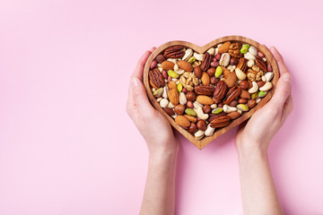 Womans hands holding heart shaped bowl with mixed nuts on pink table top view. Healthy food and...