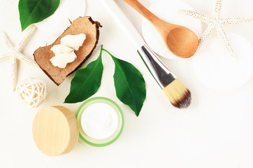 Fresh skincare moisturizer in jar with green leaves and body butter in coconut shell, view above white table cosmetic ingredients.