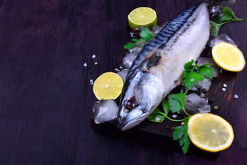 Fresh mackerel on a black chopping board surrounded by ice, lime, lemon, parsley and spices