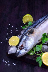 Fresh mackerel on a black chopping board surrounded by ice, lime, lemon, parsley and spices 