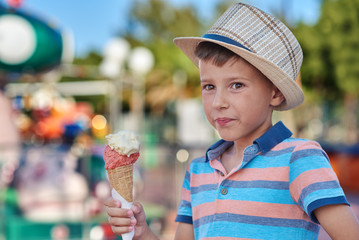 The portrait of smiling boy in a striped t-shirt and hat on his head. He is eating tasty ice-cream in the Luna park.
