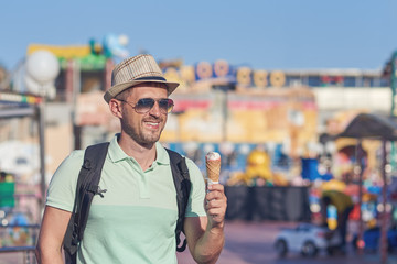 European man in the sunhat is walking around the Luna Park and eating tasty ice-cream.