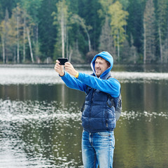 Young active European tourist is taking selfie against picturesque view on the river bank. He is enjoying his trip and smiling to the camera.