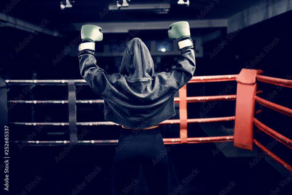 Wall mural strong caucasian boxer girl standing in ring with hands up. hoodie and boxing gloves on. backs turne