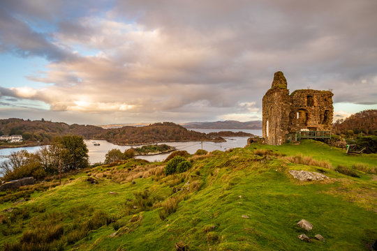 The ruins of Castle Tarbert on the right, green grass and rocks in the foreground and tarbert bay in the background with beautiful sunset clouds