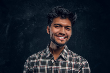 Close-up portrait of a handsome Indian man wearing a plaid shirt, smiling and looking at a camera. Studio photo against a dark textured wall