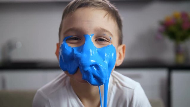 Joyful cute preadolescent child looking at camera with face covered with blue handmade slime, slowly slipping down. Funny or scary boy's face after first experiment with slime