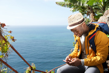 happy african american travel man sitting by sea looking at mobile phone
