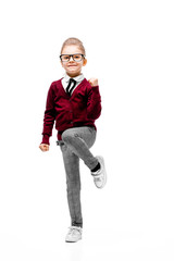 Beautiful little girl dressed like a School girl - in white shirt, red blouse and gray pants, rounded glasses standing near white wall and posing thumbs-up like model.