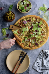 Fresh pizza with cheese, pepper and herbs on a wooden Board close-up cut and salad of greens and olives, with appliances top view on a gray stone background