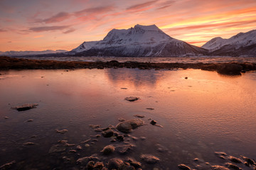 Frozen pool in foreground with beautiful reflection of snowcapped mountain in background at cloudy and colorful sunrise, Kvalöya, Norway