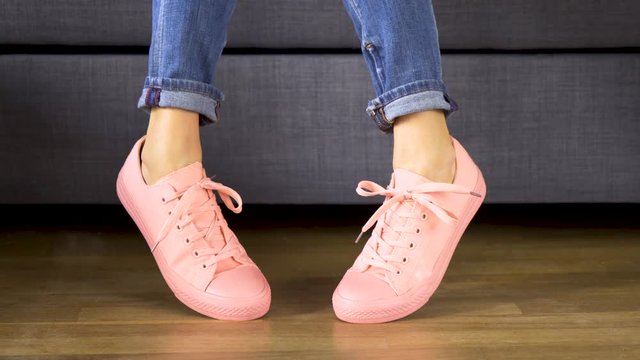 Woman is sitting on gray sofa and shows her legs in comfortable coral sneakers on wooden floor