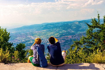 Two guys are sitting and looking into the distance and the mountains, discussing something and showing somewhere far into the distance with his hand. Sarajevo, Bosnia and Herzegovina, August 2018