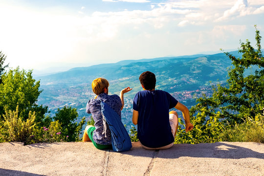 Two guys are sitting and looking into the distance and the mountains, discussing something and showing somewhere far into the distance with his hand. Sarajevo, Bosnia and Herzegovina, August 2018 - Powered by Adobe