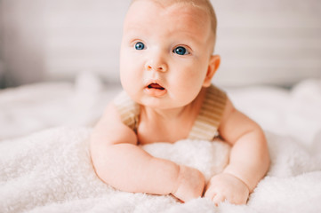 Adorable lovely newborn male baby boy with smiling emotional  happy face lifestyle indoor portrait. Funny infant child lying on stomach on bed with white wall on background.  Carefree childhood.