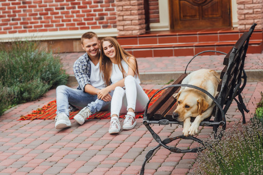 Happy young couple enjoying each other and sitting at the blanket carpet in front of the house. man and woman looks at the dog. The labradore is sleepy.