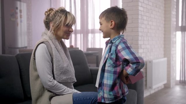 Joyful adorable little son asking mom to guess what he's holding behind back and giving gift box. Surprised with unexpected present grateful woman embracing, kissing and caressing preadolescent child