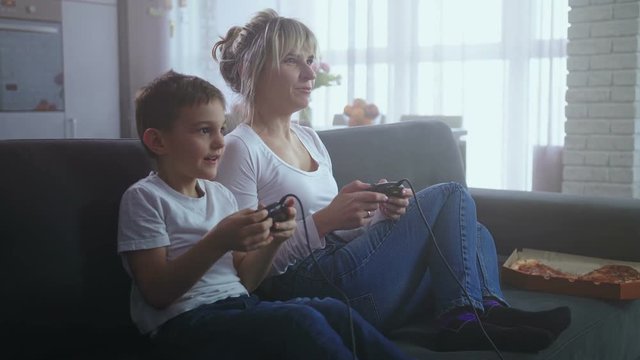 Adorable little son and pretty adult mother with joysticks playing video game sitting on couch in living room. Smiling happy family spending leisure time playing home console using game controllers