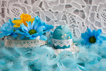 Niebieskie i żółte kwiaty, niebieskia pisanka ozdobiona białą koronką, błękitne pióra, w tle biała  koronka © Monika