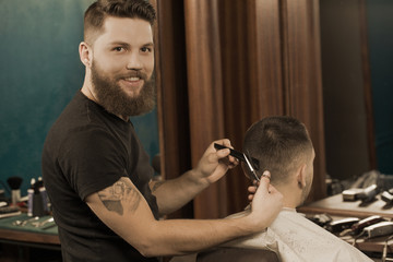 Spying on a professional barber. Handsome tattooed barber smiling happily to the camera while giving a haircut to his male client at the local barberhop