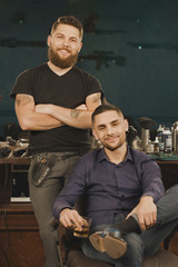 Tough men at the barbershop. Handsome male client and attractive bearded and tattooed barber posing in a barbershop