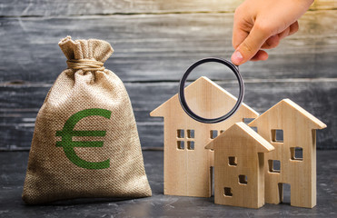 Magnifying glass is looking at a bag with euro money and three houses. Affordable cheap loan, mortgage. Taxes, rental income. Building houses. Concept of real estate acquisition and investment.