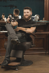 Obraz premium You will love my styling! Handsome tough and manly bearded barber sitting in a chair with a trimmer in his hand and holding a Winchester gun and smiling happily