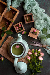 Overhead shot of composition with green teapot with tea, little wooden chest of drawers, pink roses, green gauze and old scissors on dark green concrete background