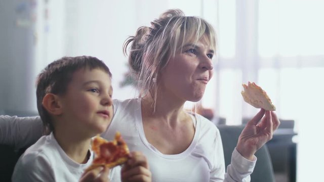 Closeup adorable little boy and adult mother having snack sitting on couch in domestic room while watching funny cartoon on TV. Eating pizza joyful family watching animation on wide TV screen