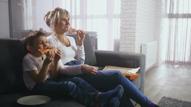 Cute preadolescent son with adult mother eating pizza and looking at TV screen showing funny child cartoon. Cheerful family having fun sitting on sofa in living room watching comic movie on TV