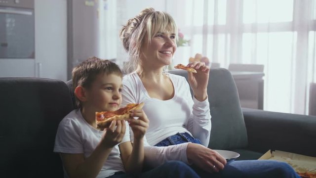 Adorable little boy and beautiful adult mother laughing while watching comedy TV show sitting on couch in domestic interior. Joyful family spending time viewing funny show on TV and eating pizza
