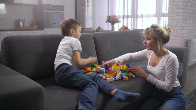 Cute little boy with adult mom deciding what toy create from plastic construction blocks scattered on sofa in domestic interior. Family spending leisure time together playing toy constructor