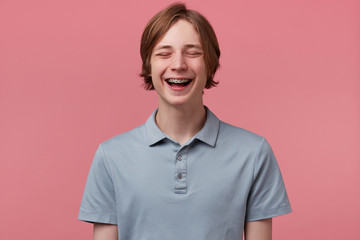 Close up of nice blue-eyed neatly combed young guy with braces on teeth joyfully laughing closed...