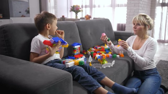 Adult mom sitting on floor and giving preadolescent boy plastic construction blocks scattered on couch. Cute little son choosing block and putting on toy constructor
