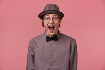 Guy opened his mouth in surprise, is overwhelmed with positive emotions happiness joy doesn't believe in his success, luck, dressed in shirt hat and black bowtie glasses has brackets isolated on pink